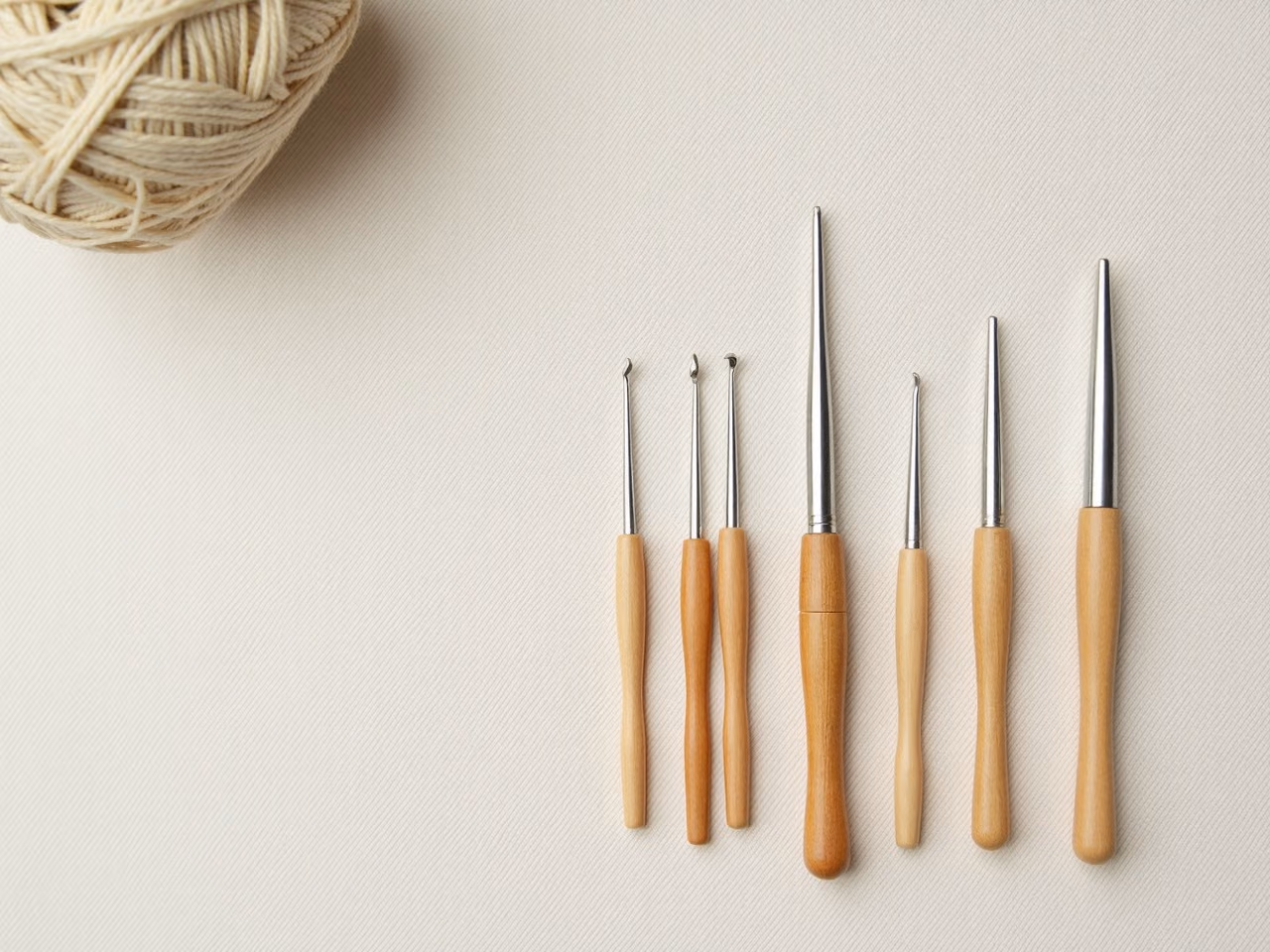 A row of crochet hooks in ascending size order laid on linen beside a ball of cream worsted yarn