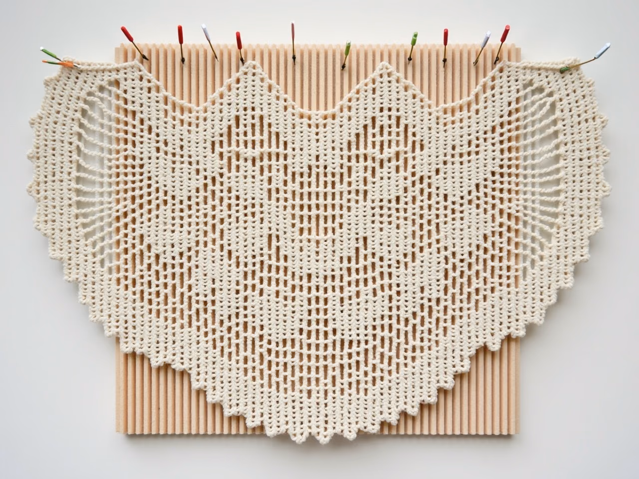 A crocheted lace shawl pinned out flat on a foam blocking mat with rustproof pins, showing the opened lace pattern